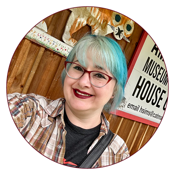 A profile photo of a woman with blue hair wearing a plaid shirt and glasses in front of a barn with cats painted on it.