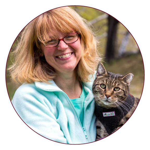 A profile photo os a blonde woman outdoors in a blue fleece jacket wearing glasses and holding a tabby cat in a harness.